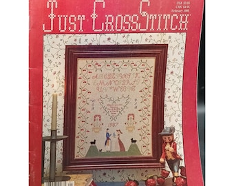 May include: A magazine cover for Just Cross Stitch, featuring a cross-stitch design of a heart with the alphabet around it, a couple in 19th-century clothing, and a small wooden figurine. The magazine cover is red with white lettering and a floral pattern.