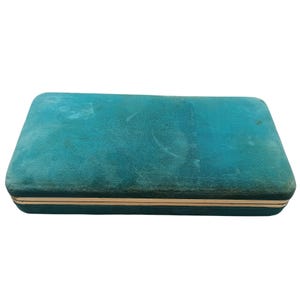 Peut inclure: Une boîte à bijoux rectangulaire, de couleur sarcelle, avec une texture semblable à du velours. La boîte a une bordure dorée sur les bords. La boîte est fermée et semble vide.