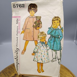 Könnte beinhalten: Vintage Simplicity-Schnittmusterumschlag mit Illustrationen von drei Kindern in Kleidern und einem Mantel. Der Umschlag ist cremefarben mit der Nummer 5762 oben. Der Text "Toddler Size 3" ist ebenfalls sichtbar.