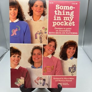 May include: A book cover titled "Something in my pocket" with designs to paint on sweatshirts. The cover shows women wearing sweatshirts with iron-on patterns, including a dinosaur, clowns, and a honey pot.