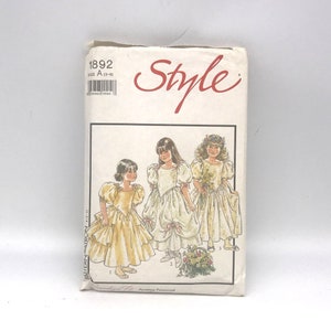 Op de afbeelding: Een vintage naaipatroonpakket met illustraties van drie jonge meisjes in formele jurken. Het pakket is gelabeld met "Style" in rood script en bevat de maatreeks "A (3-9)". Het patroonnummer is 1892.