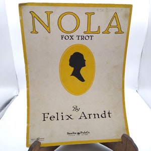 May include: A vintage sheet music cover for a song titled "NOLA" by Felix Arndt. The cover features a silhouette of a woman's head against a yellow circle with a black and white background.