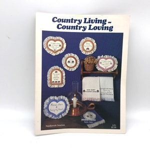 May include: A vintage needlework pattern titled "Country Living ~ Country Loving." The cover features cross-stitch designs of homes, hearts, and phrases like "Home Sweet Country Home." Includes a shelf with towels and a candle.
