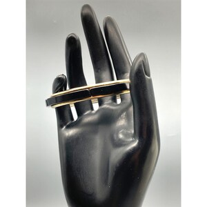 May include: A black and gold bangle bracelet with a wide, flat design. The bracelet is made of two layers of black material with a thin gold band between them.