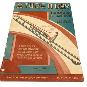 May include: A vintage music instruction book titled A Tune A Day for Trombone or Baritone by C. Paul Herfurth. The cover features a graphic illustration of a trombone against a teal and rust-colored background with text detailing its use.