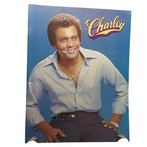 Puede incluir: Una portada de álbum vintage con un hombre sonriente con una camisa azul claro y pantalones oscuros. El nombre "Charley" está escrito en una fuente estilizada en la parte superior. El fondo es de color azul liso.