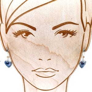 May include: Silver-toned heart-shaped earrings with a dark blue, textured design. The earrings are displayed on a wooden face-shaped jewelry holder. The earrings have a small pearl-like bead above the heart.