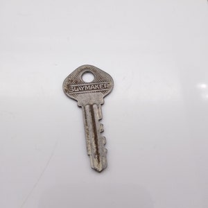 Puede incluir: Una llave de metal vintage con la palabra "SLAYMAKER" estampada en la cabeza. La llave tiene un aspecto desgastado y envejecido, con un ojo de cerradura en la parte superior y una serie de crestas a lo largo del eje. La llave es probablemente para una cerradura.