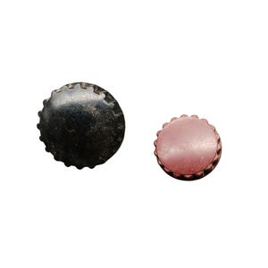 Puede incluir: Dos tapones de botella. Uno es negro con un aspecto desgastado, y el otro es de un rosa rojizo apagado. Ambos tapones tienen un diseño de borde festoneado, lo que sugiere que son para botellas.