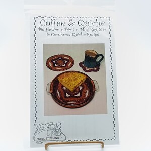 May include: A white paper pattern with a brown and orange star-shaped trivet, a mug rug, and a pie holder. The pattern also includes a recipe for cornbread quiche. The text "Coffee & Quiche" is at the top of the pattern. The text "Vogies" is at the bottom of the pattern with the text "Still Stitching..." below it.