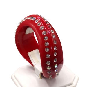 May include: A vibrant red bangle bracelet adorned with sparkling clear rhinestones. The bracelet is a bold, glossy red, with rhinestones set in two parallel rows. A striking piece of jewelry.