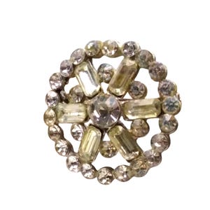 May include: A vintage, round brooch with a silver-tone metal setting. The brooch features a central, large, clear, round rhinestone surrounded by a ring of smaller, clear rhinestones. Rectangular and round rhinestones are arranged in a starburst pattern.