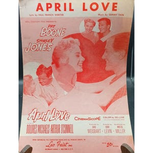 Pode incluir: Partitura de "April Love" com Pat Boone e Shirley Jones. A capa é predominantemente vermelha e branca, com imagens dos atores e o título do filme. O texto inclui os nomes do letrista, compositor e créditos de produção.