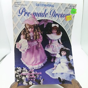 May include: A book cover with the title "Premade Dresses" for 13 to 16 inch porcelain-look dolls. The cover features three dolls wearing different dresses. The dolls are wearing pink, blue, and white dresses. The book cover is decorated with a white lattice pattern.