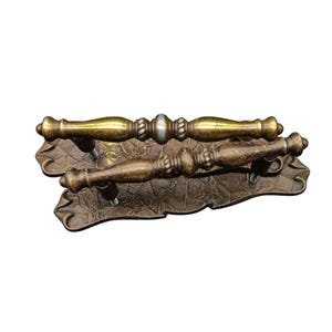 May include: Two metal drawer pulls with a brass finish, featuring a central decorative band and a textured, dark bronze-toned backplate with an ornate, scalloped edge. The handles have a smooth, rounded profile with subtle rope-like detailing.