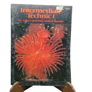 May include: A music instruction book titled Intermediate Technic 1 by James and Jane Smisor Bastien. The cover features a black background with a large, vibrant orange sea anemone-like graphic and white text at the bottom.