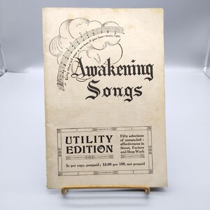 May include: Vintage "Awakening Songs" booklet. The cover features the title in ornate black lettering, with musical notes and a cloud illustration. The "Utility Edition" is also displayed. The booklet is propped up on a wooden stand.