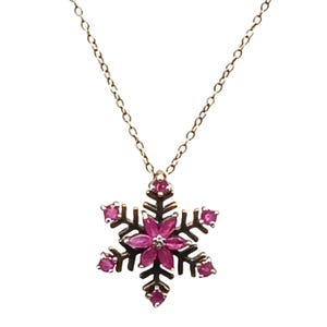 May include: A gold-toned necklace featuring a snowflake pendant. The pendant has a dark metal frame with a central flower design and six outer points, each adorned with a vibrant pink gemstone. The necklace chain is delicate.