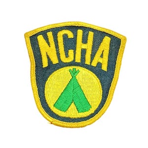 May include: A shield-shaped embroidered patch with a dark blue background and a yellow border. The text NCHA is stitched in yellow at the top. Below the text, a yellow circle contains a green graphic of a teepee with a small flag on top.
