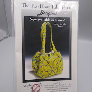 Può includere: Una borsa tote in tessuto con stampa floreale gialla e viola con fodera blu. La borsa ha un'unica tracolla e una piccola chiusura a bottone blu. Il testo sull'immagine recita "The Two-Hour Tulip Purse Bouquet Now available in 4 sizes! Larger size takes longer."