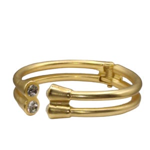 May include: A gold-colored open cuff bracelet with two clear, round gemstones on one end. The bracelet has a hinge and a double-band design. The metal has a matte finish.