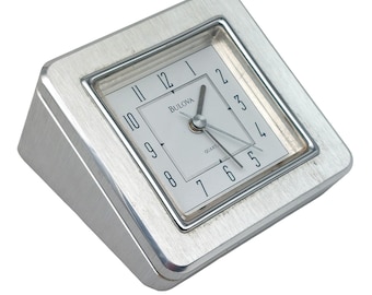 Può includere: Un orologio quadrato argentato con un quadrante bianco e lancette nere. L'orologio dice "Bulova Quartz" sul quadrante.