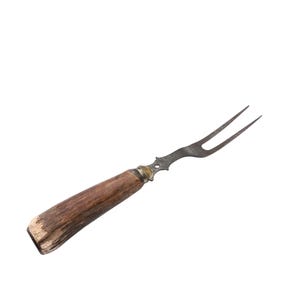 Op de afbeelding: Een vintage trancheervork met een metalen kop met twee tanden en een bruine, getextureerde handgreep. De handgreep heeft een messing kraag en een versleten, rustieke uitstraling. De vork staat tegen een effen witte achtergrond.