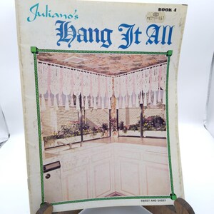 May include: Vintage book cover titled "Juliano's Hang It All" in teal script and ornate font. The cover features a photo of a kitchen with lace curtains and a floral ceiling. The book is labeled "Book 4" and "Sweet and Sassy".