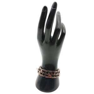 May include: A copper-coloured bracelet featuring a series of connected circular links is displayed on a black hand-shaped jewellery holder. The bracelet has a polished finish and a vintage aesthetic. The background is plain white.