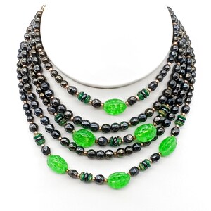 Op de afbeelding: Een meerrijige ketting met zwarte kralen en groene glazen kralen. De ketting is gelaagd en creëert een cascade-effect.
