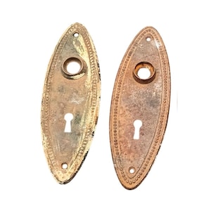 Könnte beinhalten: Zwei ovale antike Türschilder mit Schlüssellochöffnungen. Die Metallplatten haben ein abgenutztes, gealtertes Aussehen. Eine Platte hat eine goldene Oberfläche, die andere eine bronzefarbene. Beide haben dekorative Perlenränder und kleine Schraubenlöcher.