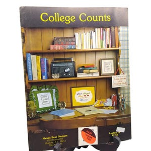 May include: A craft pattern leaflet titled College Counts featuring a wooden hutch with shelves holding books, a radio, and framed cross-stitch designs. The desk surface displays a green pillow, a yellow framed sign, papers, and a pen.