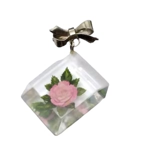 May include: A clear glass pendant featuring a pink rose and green leaves. The pendant is attached to a silver bow.