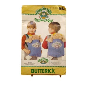 May include: A vintage Butterick sewing pattern featuring Cabbage Patch Kids. The pattern packet is yellow with images of children wearing denim backpacks with plush doll heads. Text includes "Cabbage Patch Kids" and "P'tits Bouts de Choux".