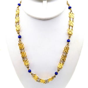 May include: A gold-toned necklace with intricate openwork floral designs and interspersed with small, round, deep blue beads. The necklace is displayed on a white mannequin bust, showcasing its length and design.