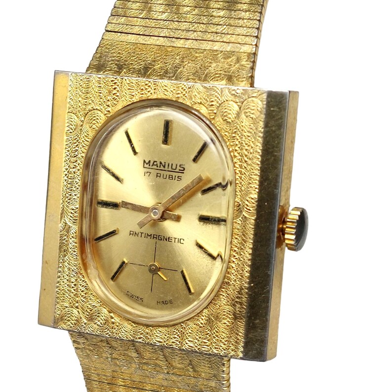 Puede incluir: Un reloj de pulsera dorado con una caja cuadrada y una esfera de forma ovalada. La esfera tiene marcadores de hora romanos negros, agujas negras y el texto "MANIUS 17 RUBIS" y "ANTIMAGNETIC".