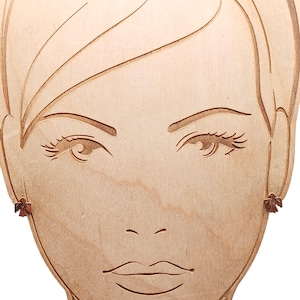 Pode incluir: Expositor de madeira com um rosto esculpido e brincos. Os brincos são pequenos, castanho-avermelhados e em forma de pássaros. O expositor é feito de madeira clara, com as feições do rosto gravadas.