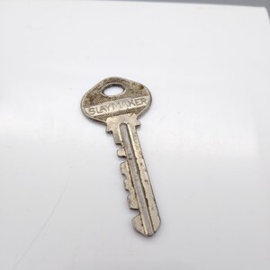 Puede incluir: Una llave plateada con la palabra "SLAYMAKER" estampada en la cabeza. La llave tiene un agujero circular en la parte superior y una serie de muescas a lo largo del eje. La llave está sobre un fondo blanco.
