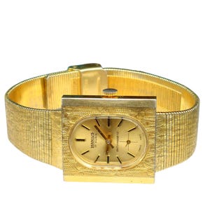 Puede incluir: Un reloj de pulsera dorado con una esfera cuadrada y una correa de metal dorado. La esfera del reloj tiene una esfera dorada con agujas negras y marcadores negros. La esfera del reloj tambi&eacute;n tiene el texto "MANIUS 17 RUBIS ANTI-MAGNETIC" impreso en ella.