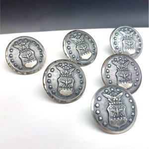 Puede incluir: Un conjunto de siete botones metálicos plateados. Cada botón presenta un emblema detallado con un águila, estrellas y las letras "AF". Los botones tienen una apariencia ligeramente envejecida, sugiriendo un estilo vintage.