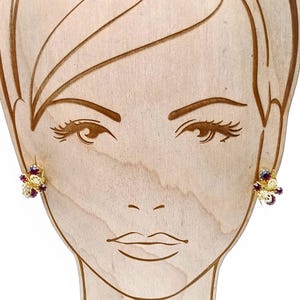 May include: A pair of gold earrings with red gemstone accents. The earrings are in the shape of a flower with a delicate design.