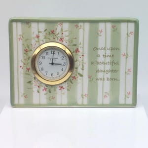 May include: A rectangular ceramic clock with a gold-colored clock face. The clock face has black numbers and hands. The clock is decorated with a green and white striped pattern and floral accents. The text "Once upon a time a beautiful daughter was born" is printed on the clock.