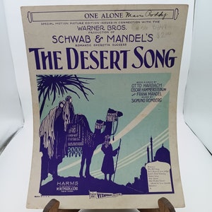 Puede incluir: Portada de partitura de "The Desert Song", una opereta romántica. La portada presenta una ilustración púrpura de un camello con jinetes y una figura sosteniendo un bastón, sobre un fondo desértico. El texto incluye el título, detalles de producción y créditos de los compositores.
