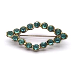 Puede incluir: Broche ovalada con un marco de metal dorado. El marco está adornado con piedras preciosas redondas de color verde azulado. La broche tiene un alfiler en la parte posterior para sujetarla. Una joya de estilo vintage.