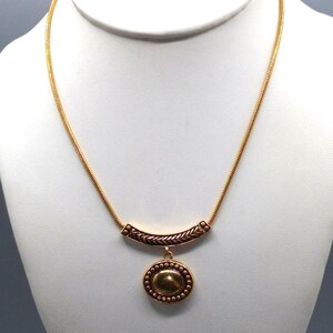 Könnte beinhalten: Eine goldfarbene Halskette mit einer filigranen Kette. Die Halskette hat einen gebogenen Barrenanhänger mit strukturiertem Design und einen runden Anhänger mit Perlenrand und einem ovalen Mittelelement. Der Schmuck wird auf einer weißen Schaufensterpuppe präsentiert.