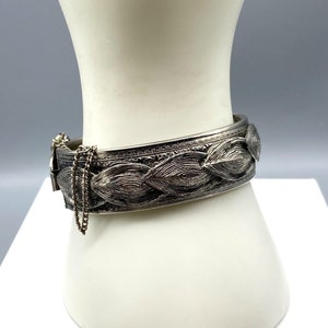 May include: A silver tone hinged bangle bracelet with a leaf design. The bracelet has a chain attached to the clasp.