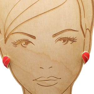 May include: A pair of stud earrings featuring a teardrop shape with a vibrant red enamel finish and a thin gold-toned metal outline. The earrings are displayed on a light-colored wooden mannequin head cutout that shows a stylized facial outline.