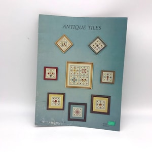 Op de afbeelding: Een boekomslag met de titel "Antique Tiles" met een blauwe achtergrond en afbeeldingen van ingelijste kruissteekontwerpen. De boekomslag bevat ook de tekst "€ 5,00 BOEK 1".