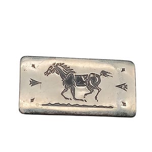 May include: A rectangular silver-toned metal belt buckle featuring a dark engraved design of a galloping horse in the center. The design includes small star and arrow motifs on either side of the horse, with a horizontal line beneath the animal.