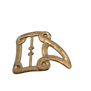 Puede incluir: Hebilla de cinturón vintage de color latón con un diseño abstracto. La hebilla presenta un marco rectangular con una barra vertical central y un elemento curvo y alargado. El metal tiene un aspecto ligeramente envejecido.